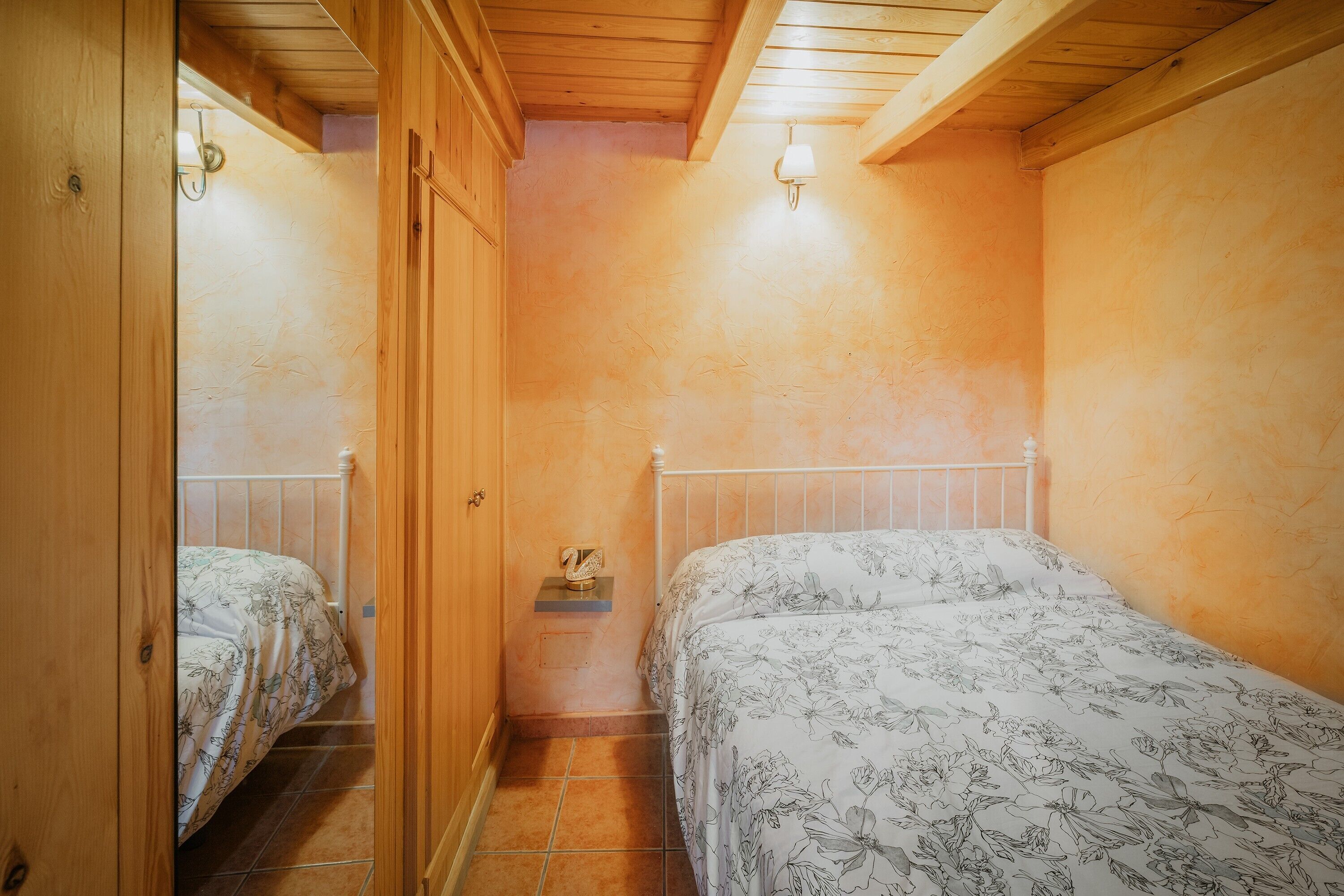 A bedroom with two beds, wooden walls, a tiled floor, and a small bedside table with a lamp.