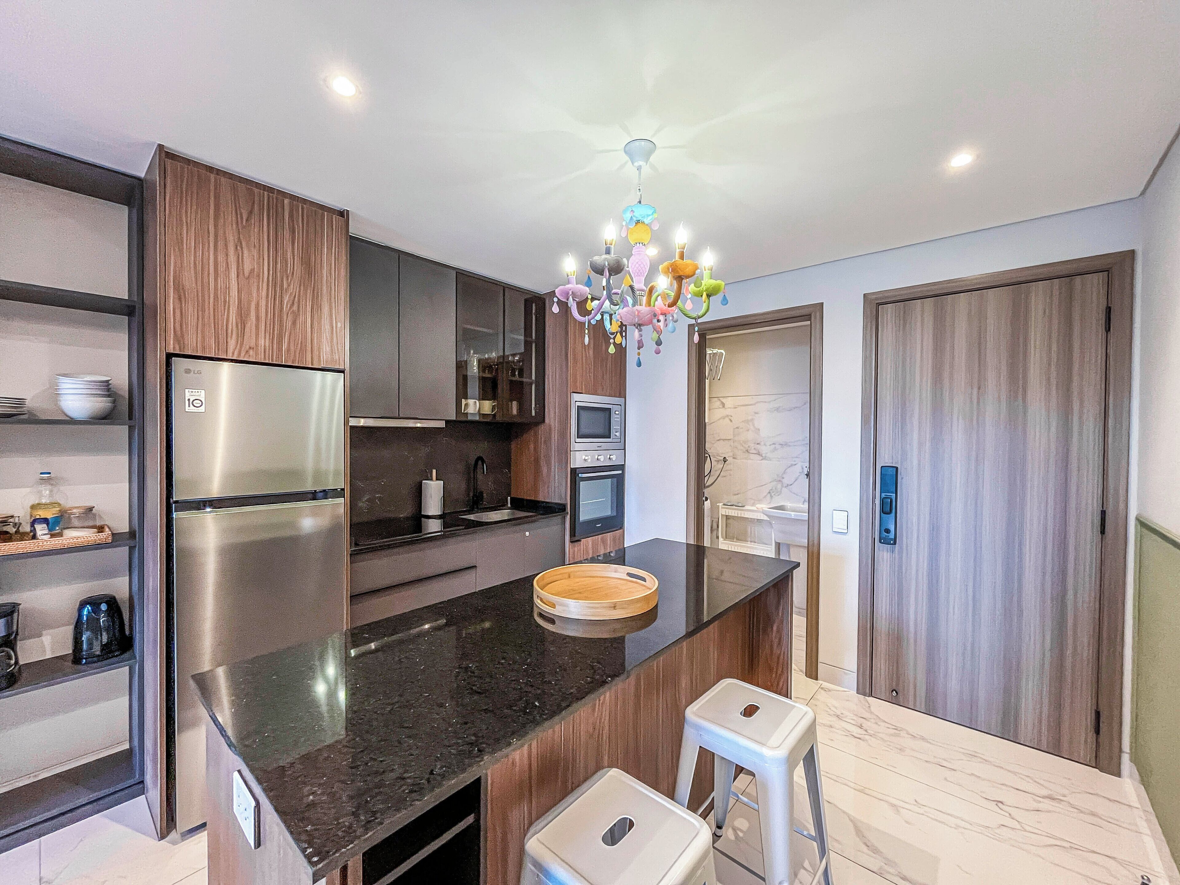 Uma cozinha moderna com geladeira de aço inoxidável, bancadas escuras, armários de madeira, micro-ondas e um lustre.