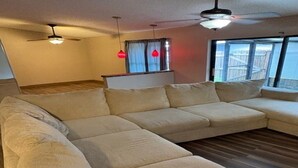 A modern living room with a large beige sectional sofa, a ceiling fan, and a window with curtains. - Stay Where Tampa Comes Alive - Central Home Near Events & Attractions (Tampa)