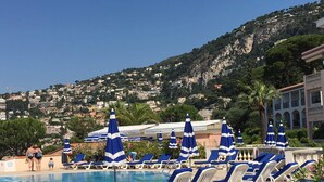 A pool area with blue and white sun loungers and umbrellas, a clear view of a hillside with buildings, and a palm tree. - Apartment Ange Gardien, swimming pool, panoramic sea view in Villefranche sur Mer (Villefranche-sur-Mer)
