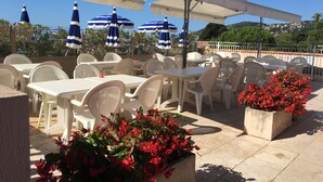 An outdoor seating area with white plastic chairs and tables, blue-striped umbrellas, and red flowers in planters. - Apartment Ange Gardien, swimming pool, panoramic sea view in Villefranche sur Mer (Villefranche-sur-Mer)