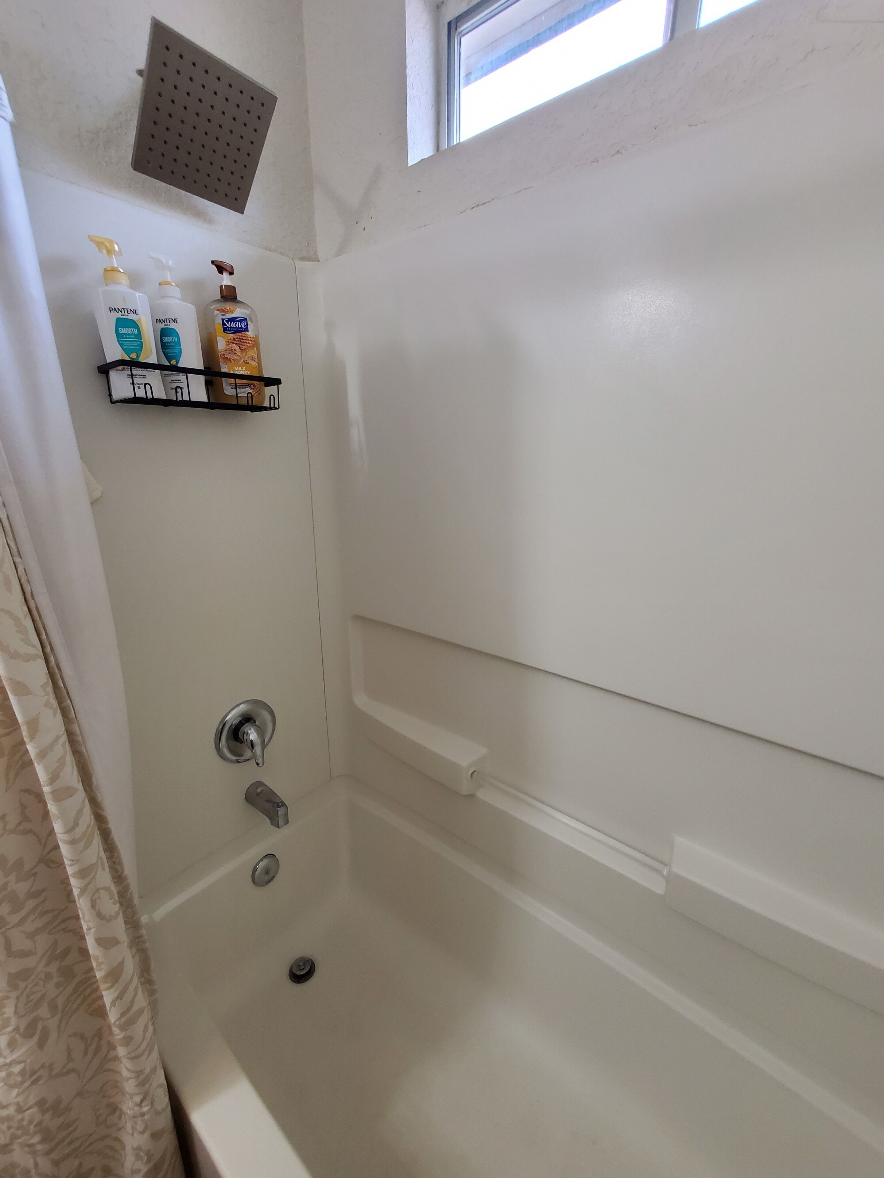 A bathroom with a bathtub, a showerhead, a window, and a shelf with toiletries.