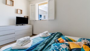 Une chambre à coucher avec un lit, un téléviseur posé sur une commode, une fenêtre avec des volets ouverts et une vue sur un bâtiment à l’extérieur.