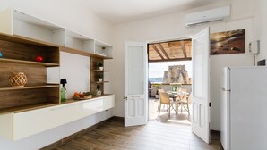 Une cuisine moderne avec des étagères en bois, un réfrigérateur blanc et une vue sur la mer, visible à travers une porte ouverte.
