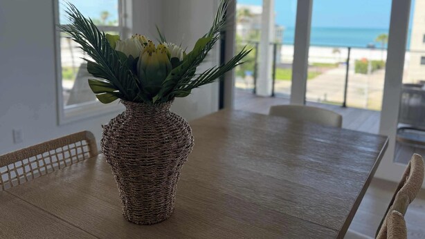 Ett matbord med en flätad vas full av blommor, utsikt över havet genom stora fönster och en balkong med ett räcke.