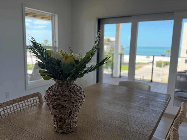 A dining table with a woven vase of flowers, a view of the ocean through large windows, and a balcony with a railing. - Chateau Bleu ·2026 Luxury· Rooftop Deck Gulf View (Fort Myers Beach)