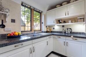 A kitchen with white cabinets, a granite countertop, a fruit bowl, and a window with a view of greenery. - Authentic & Stylish Gem in the Heart of Chipping Campden: Toys, Books, Workspace (Chipping Campden)