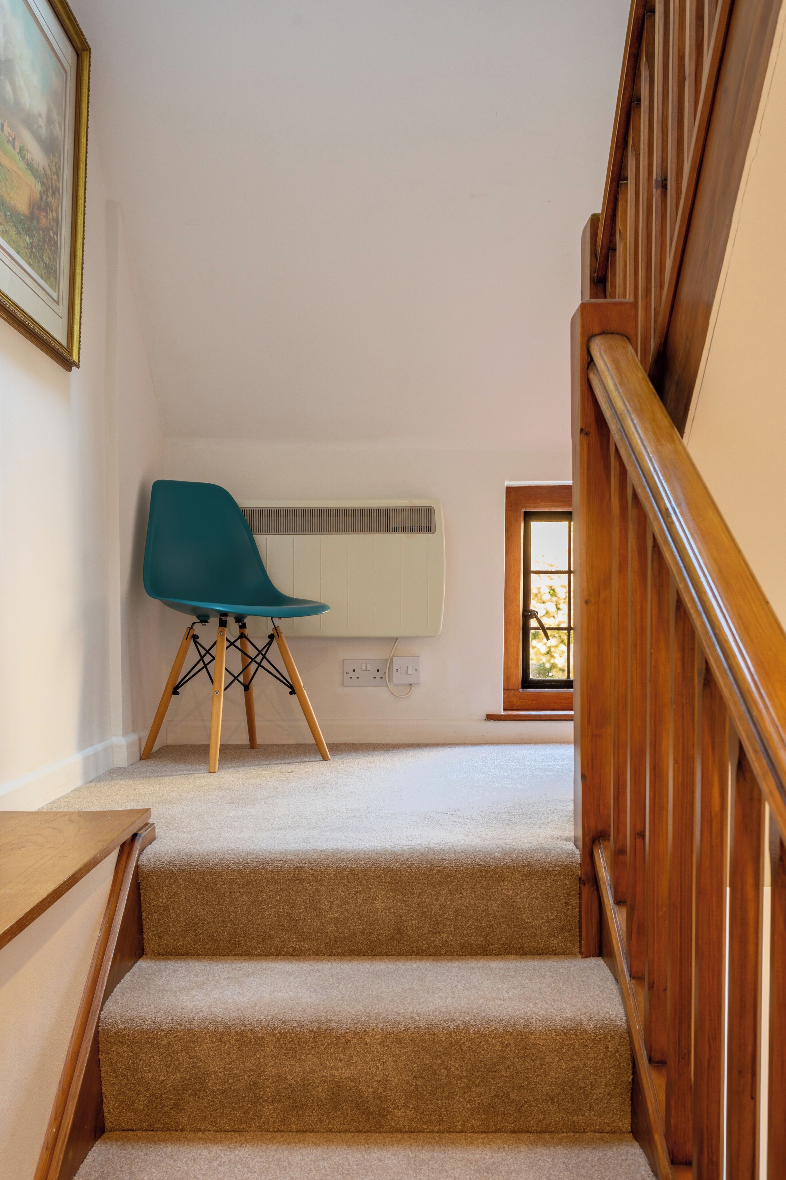 Un escalier dont les marches sont recouvertes de moquette mène à un palier. Une chaise de couleur turquoise est placée sur le palier, et un radiateur blanc est fixé au mur.