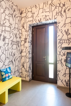 Una habitación con un papel tapiz estampado, una puerta de madera, un banco amarillo y un almohadón.