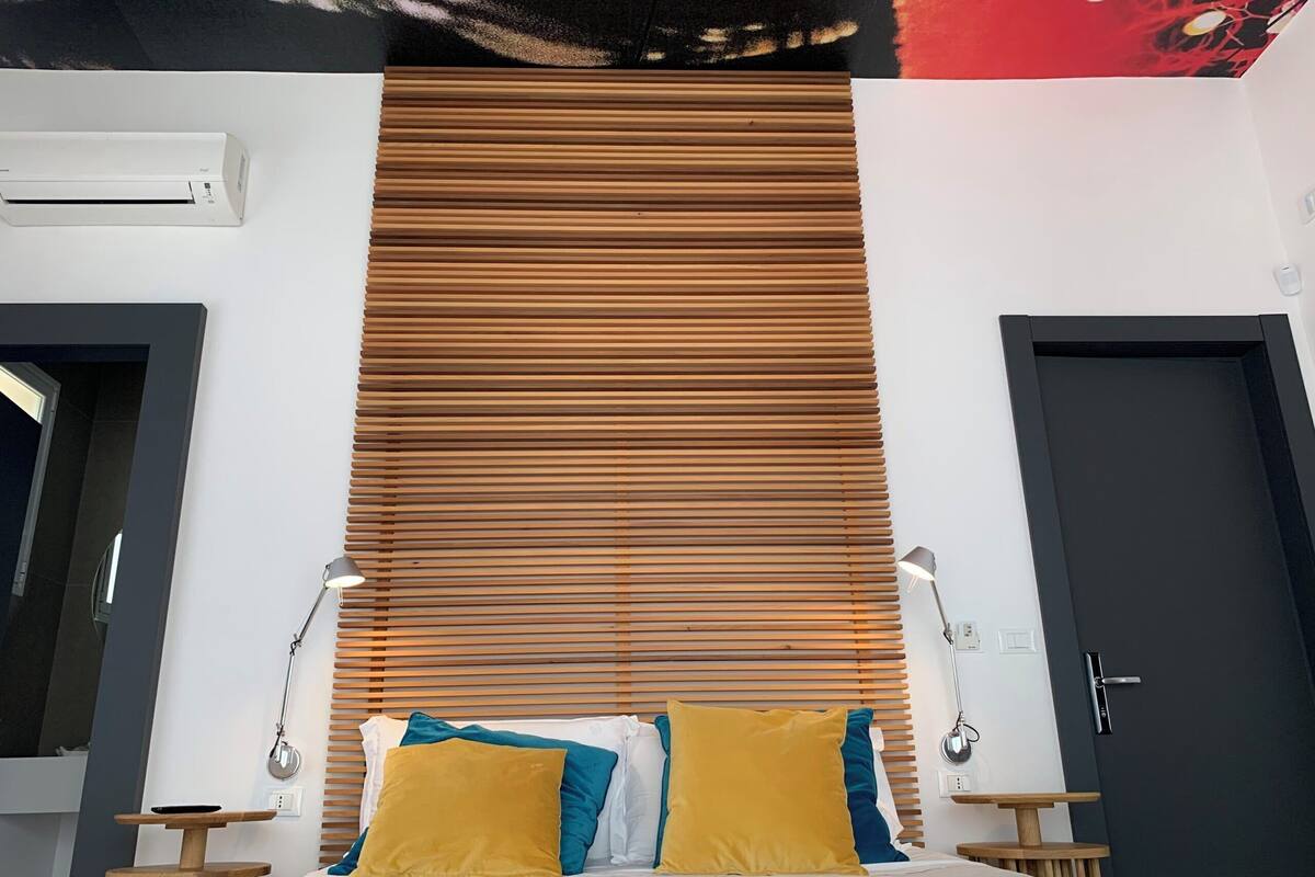 Une chambre d’hôtel moderne, équipée d’un lit, de stores en bois, de deux tables de chevet avec des lampes et d’une porte.