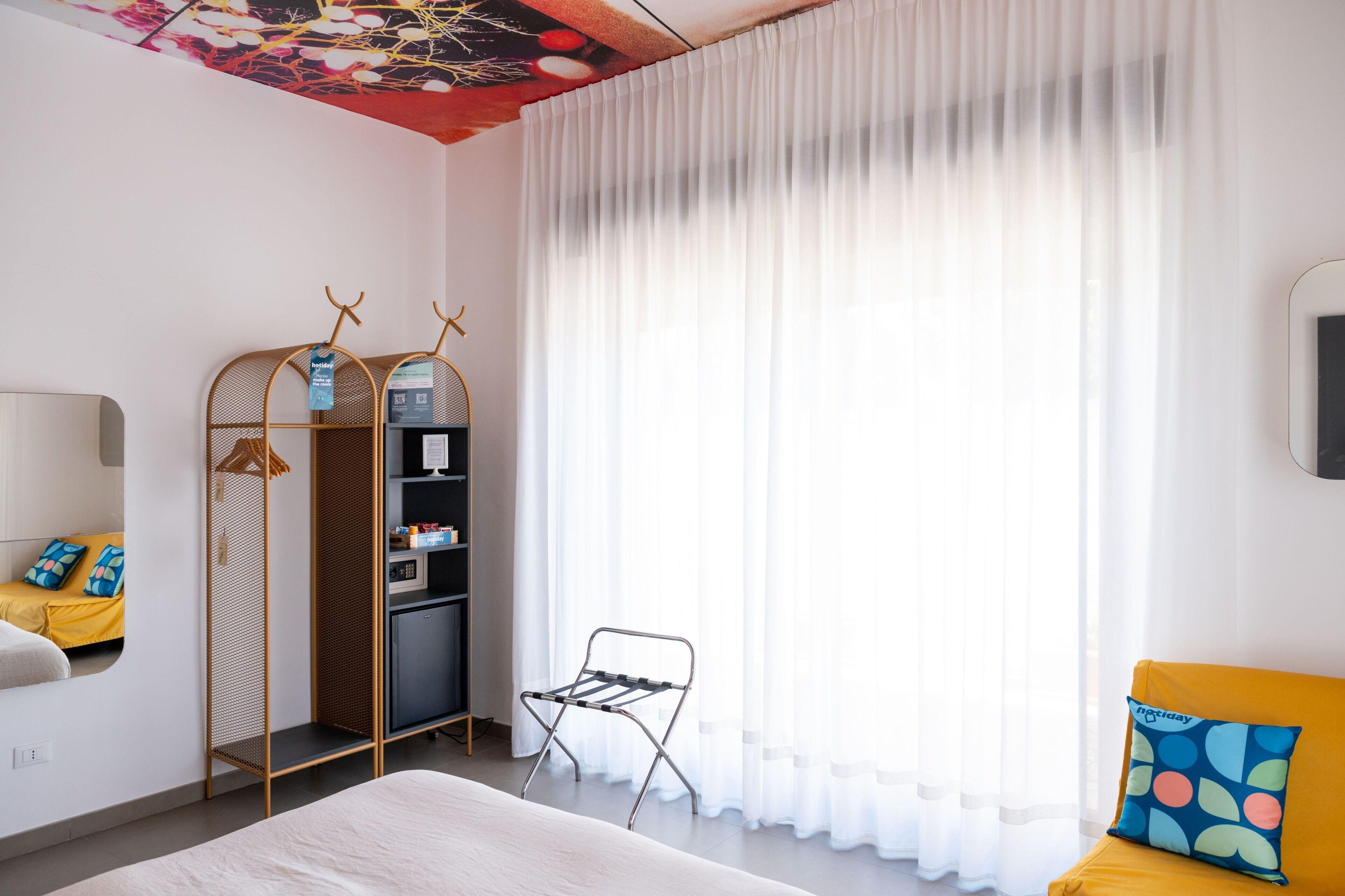 Ein modernes Hotelzimmer mit einem Bett, einer Sitzecke mit einem gelben Sofa, einem kleinen Tisch, einem Wäscheständer aus Metall und einem Kleiderschrank.