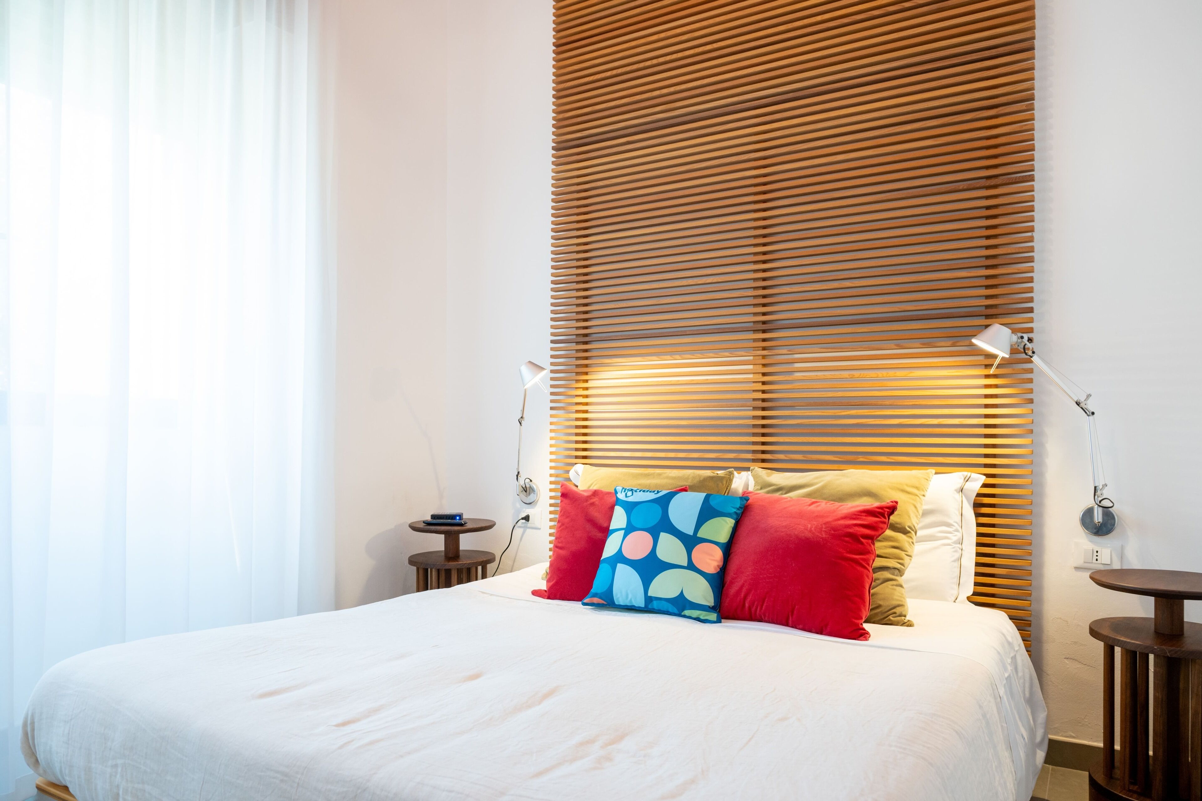A neatly made bed with white bedding and colorful pillows, a wooden slatted headboard, bedside tables with lamps, and a window with sheer curtains.