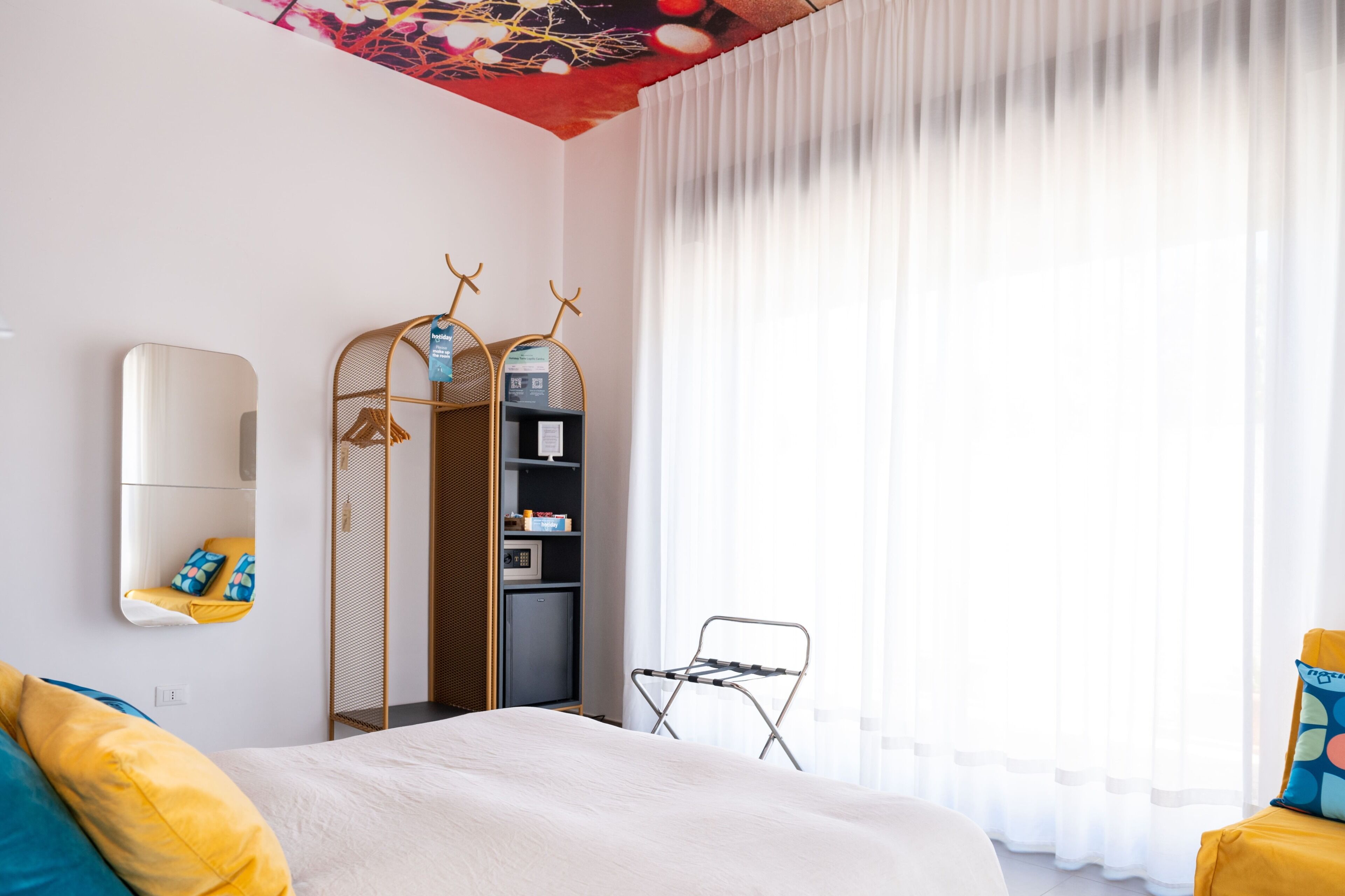 Ein modernes Hotelzimmer mit Bett, Kleiderschrank, Spiegel und einem Fenster mit durchsichtigen Vorhängen.