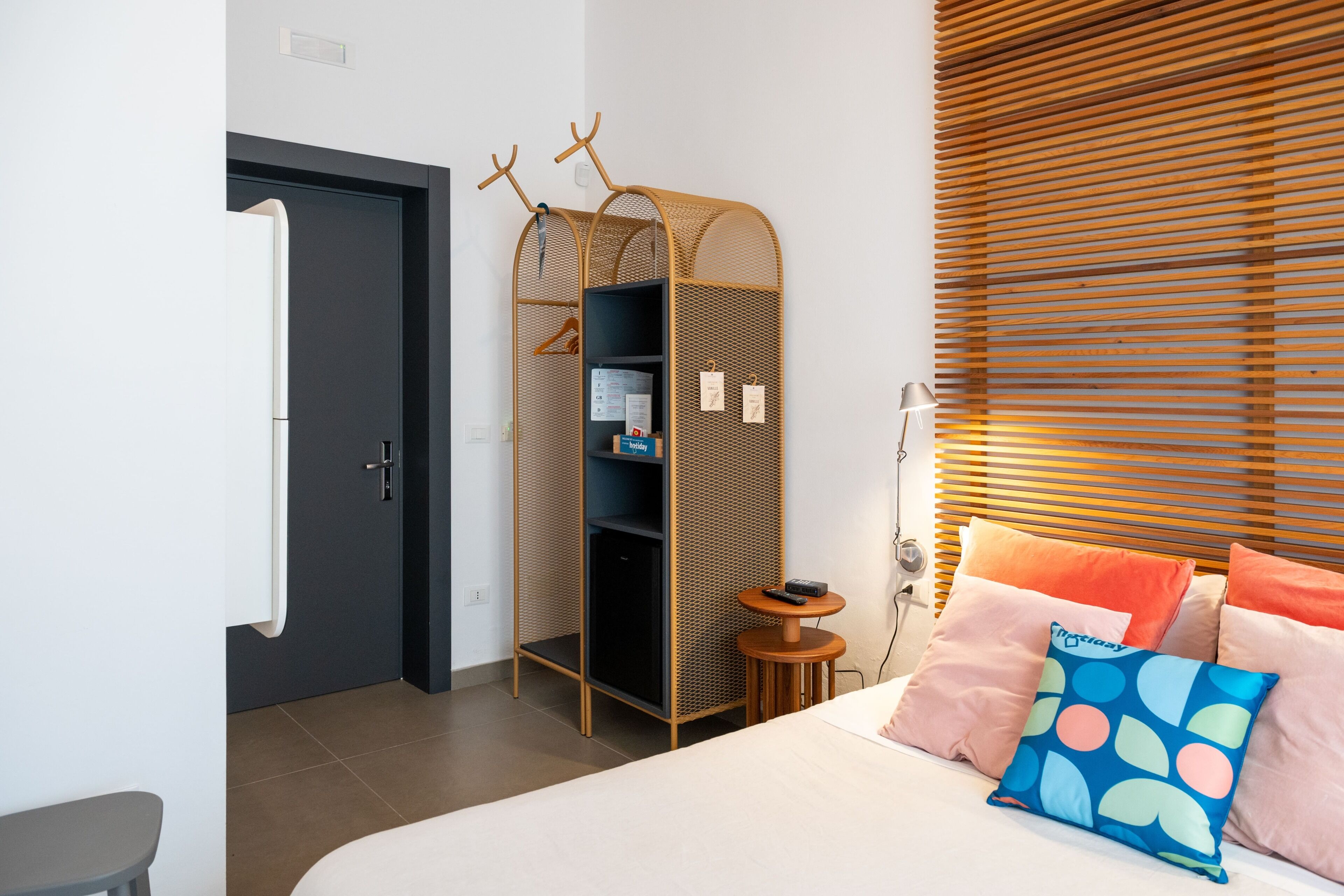 A modern hotel room with a bed, a wardrobe with a mirror, a small table, and a window with blinds.