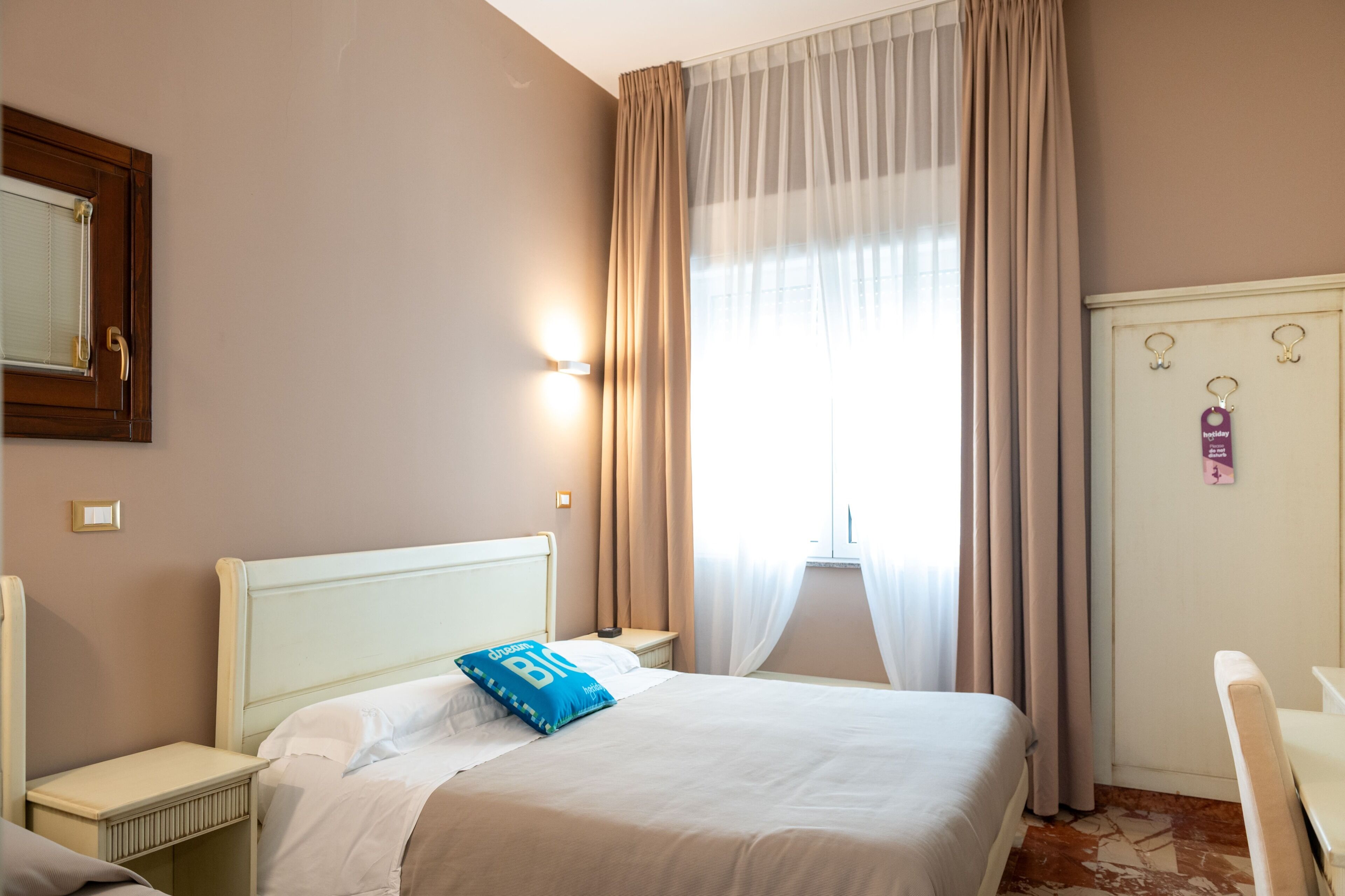 Ein Hotelzimmer mit Bett, Nachttisch, Schreibtisch, Kleiderschrank und einem fensterbepflanzten Fenster.