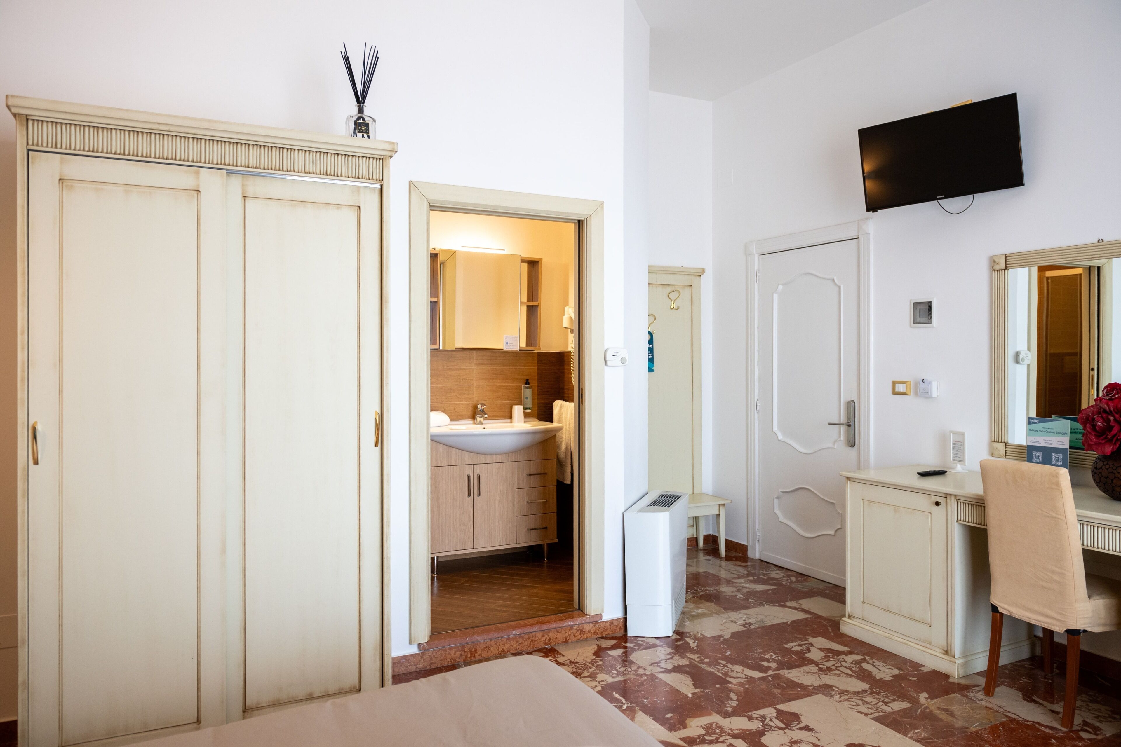 Une chambre comprenant un lit, une table de chevet, un téléviseur fixé au mur, un miroir et une porte donnant sur une salle de bain équipée d’un lavabo et de placards.