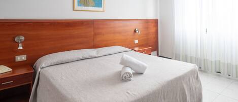 Una cama bien hecha con una colcha blanca, un cabecero de madera, una mesita de noche con una lámpara y un cuadro en la pared.