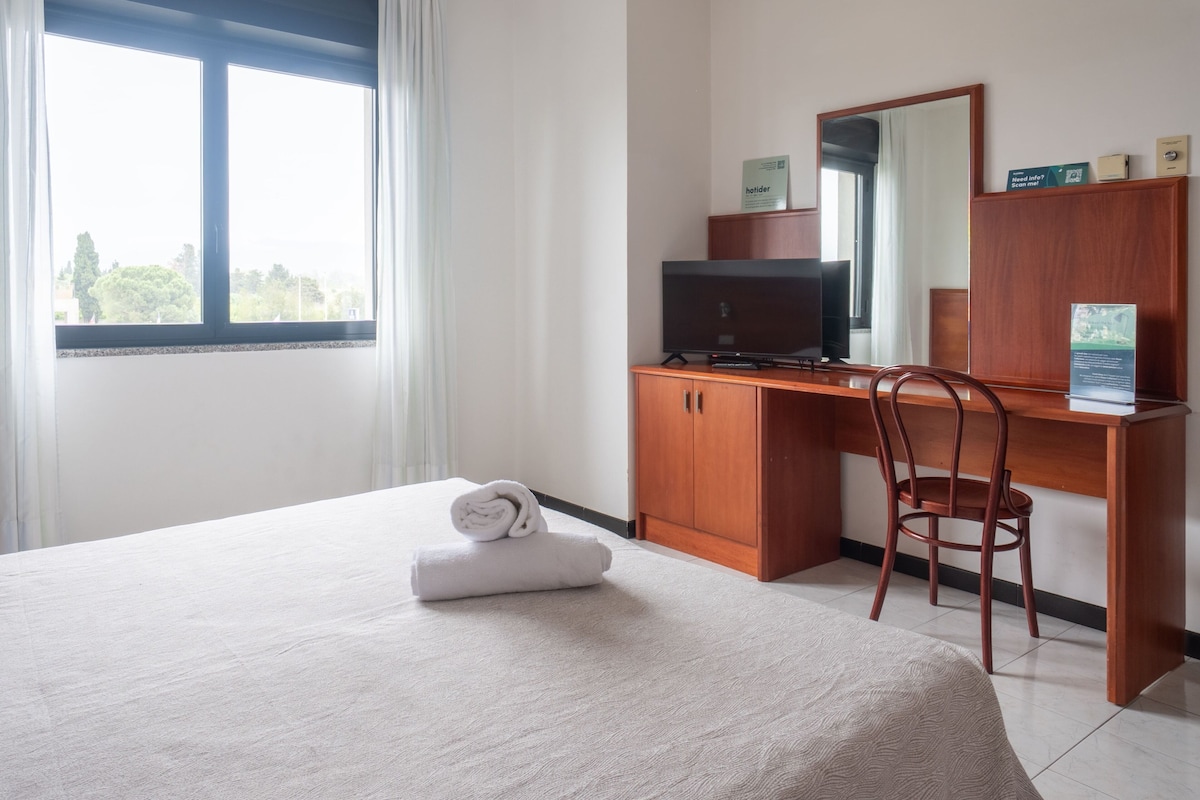 A hotel room with a bed, a desk with a chair, a television, and a window with curtains.