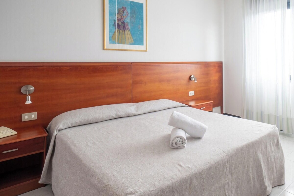 A hotel room with a wooden headboard, a bed with a grey bedspread, a rolled towel on the bed, a nightstand with a lamp, and a painting on the wall.