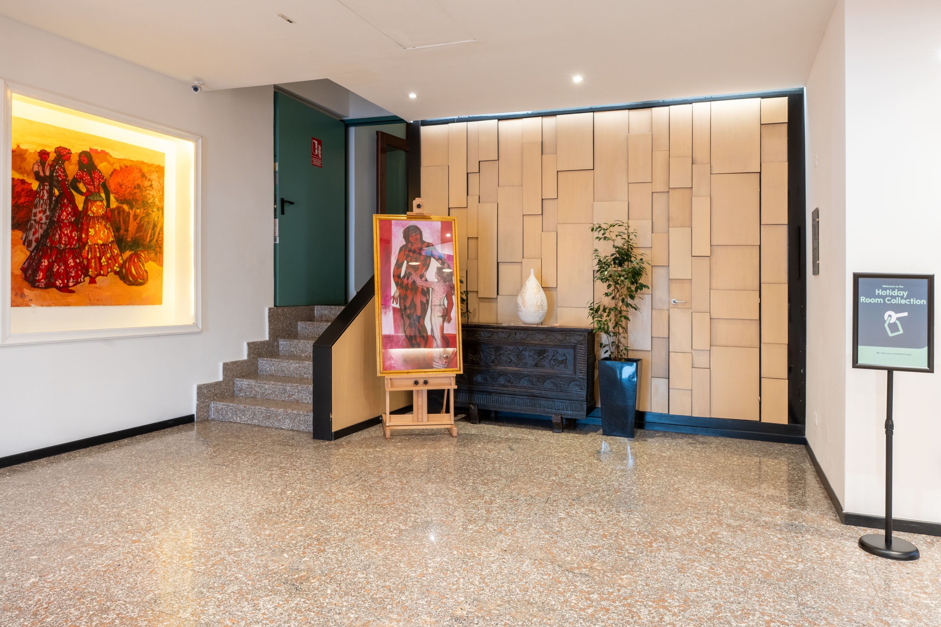 A lobby with a staircase, a large painting, a decorative wall, and a sign indicating a holiday room collection.