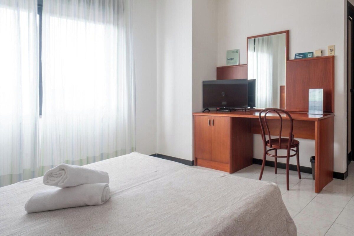 A hotel room with a bed, a desk with a computer, a chair, and a window with sheer curtains.