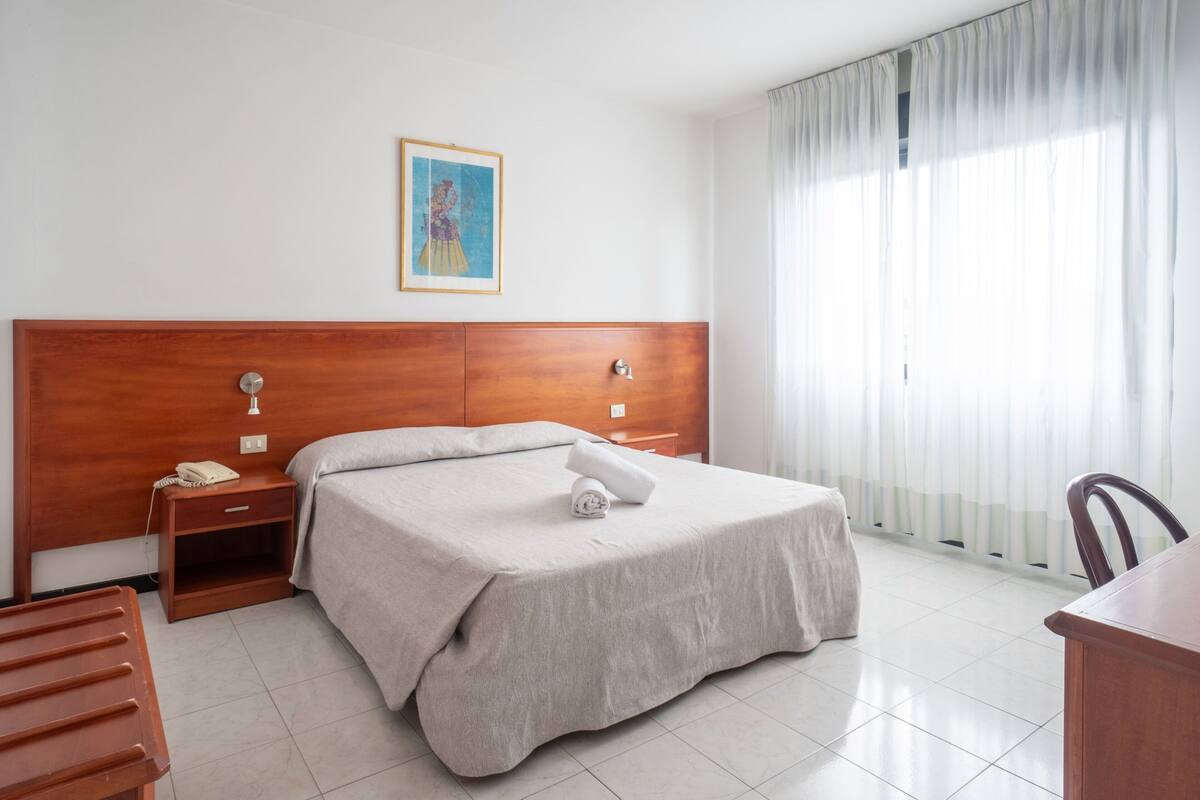 A hotel room with a wooden headboard, a bed with a beige bedspread, a bedside table with a phone, a chair, and a large window with curtains.