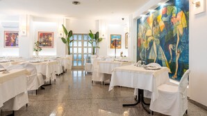 A restaurant with tables set for dining, a large abstract painting on the wall, and a plant in the center. - Hotiday Cagliari Sant'Elena (Quartu Sant'Elena)