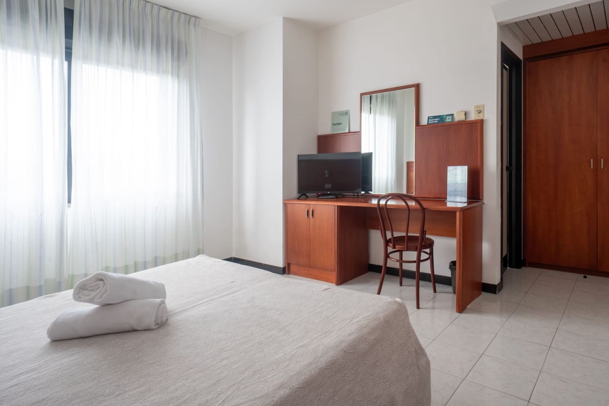 A hotel room with a bed, a desk with a computer, a chair, a wardrobe, and a window with curtains.