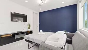 A modern room with a grey sofa, a small table, and a flat-screen TV mounted on the wall. - Seaglass Retreat! Pool Escape, Walk to Beach + DT Lauderdale by the Sea (Lauderdale-by-the-Sea)