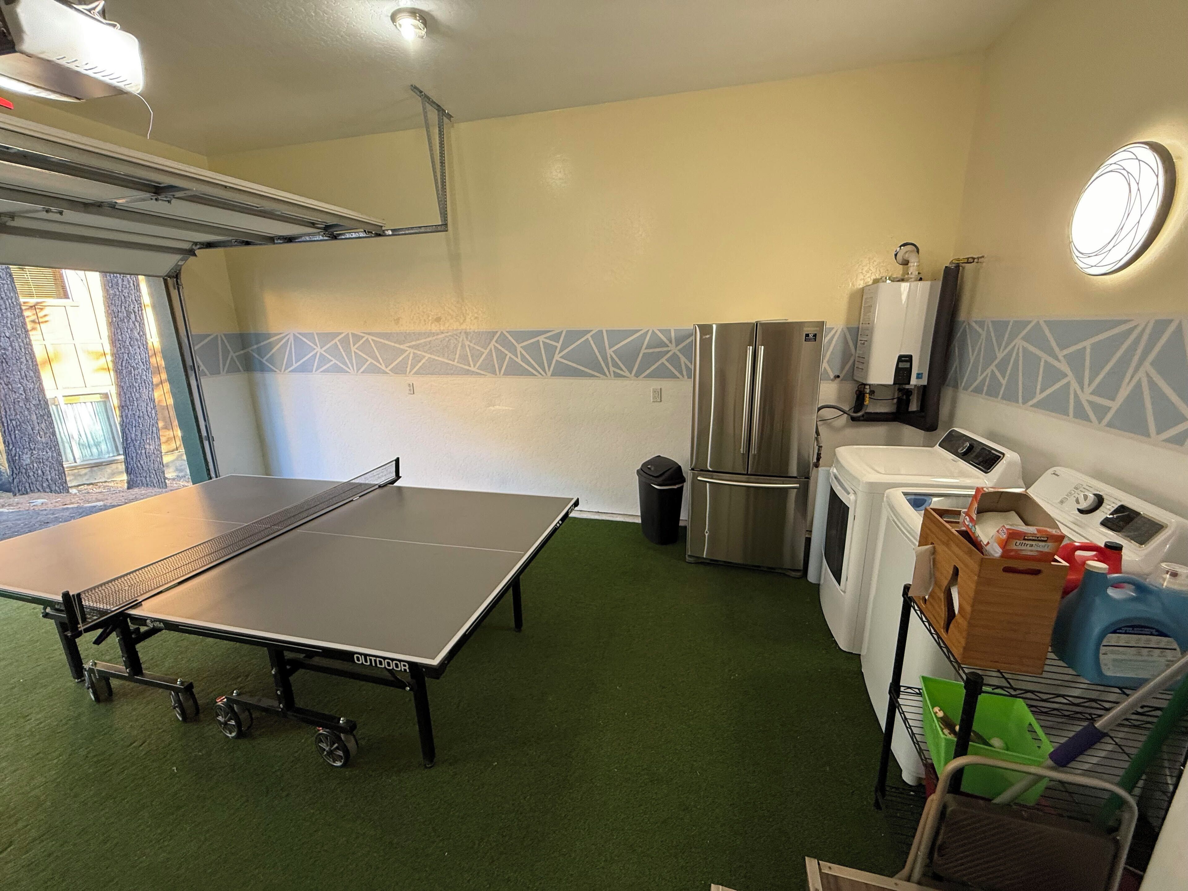 A compact kitchen with a ping pong table, a stainless steel refrigerator, a washing machine, and a microwave.