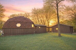 A barn with a hexagonal skylight, surrounded by trees and a grassy field. - Lakefront Dome Retreat (Maple Lake)