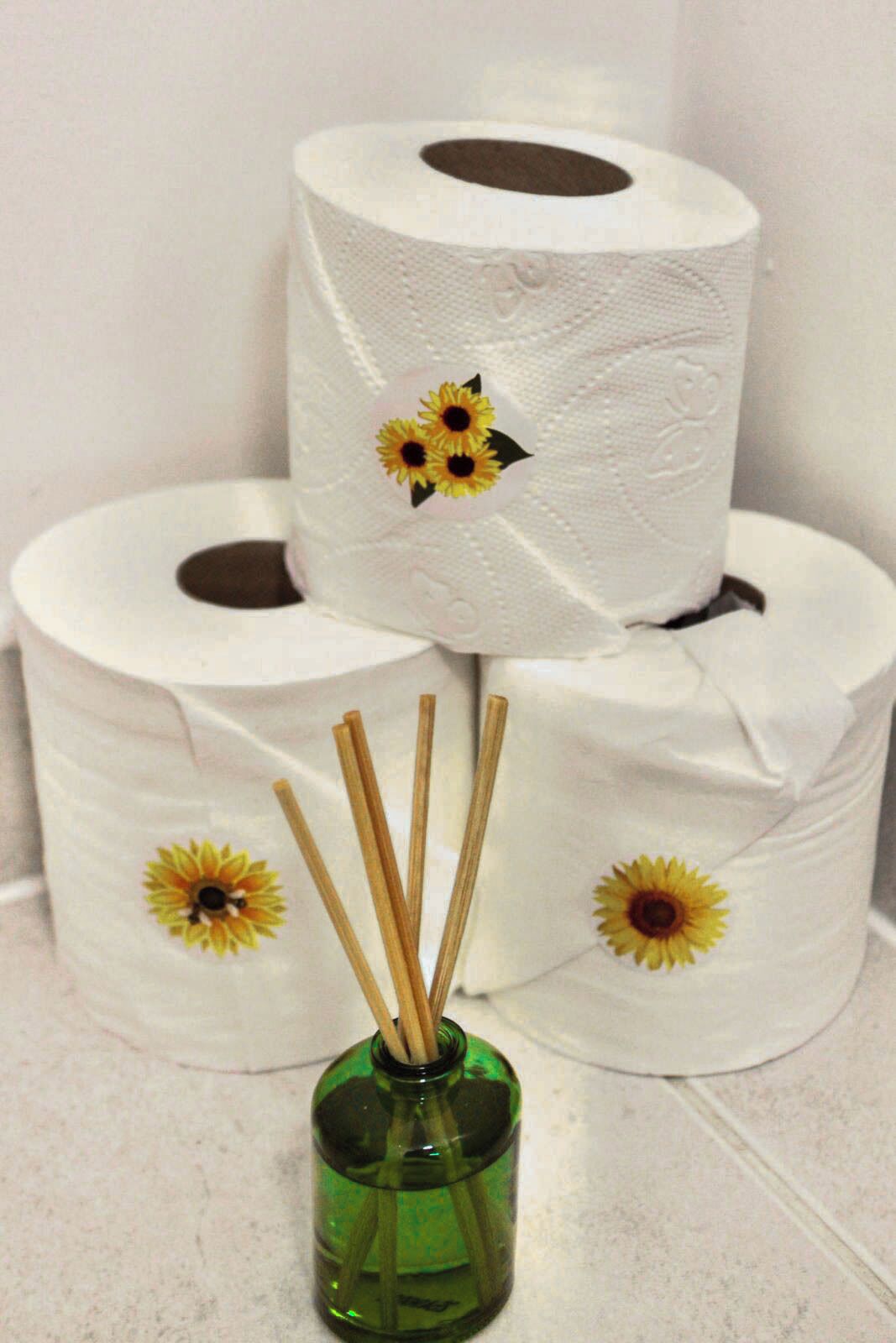 Trois rouleaux de papier toilette ornés de motifs de tournesols, un vase en verre vert avec des baguettes en bois, et un sol carrelé.