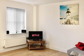 A room with a wooden TV stand, a white radiator, a window with blinds, and a painting of a lighthouse.
