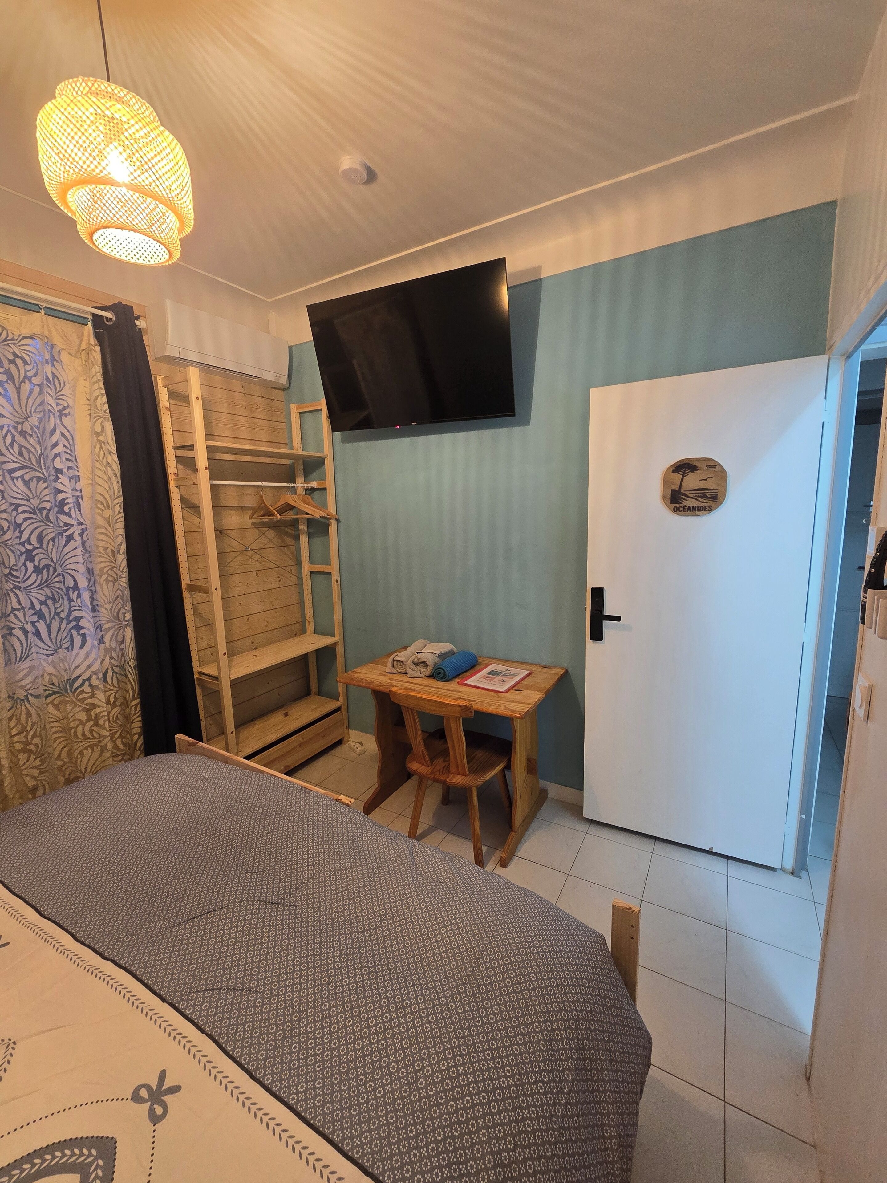 A bedroom with a bed, a TV mounted on the wall, a wooden table with a chair, and a wardrobe.