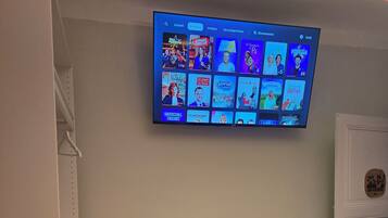 Habitación de hotel con un televisor fijado a la pared mostrando varios canales. Hay un pequeño escritorio blanco con un cojín a rayas rojas y blancas, un taburete rojo y un libro encima. La habitación tiene un revestimiento de pared de madera y una puerta blanca.