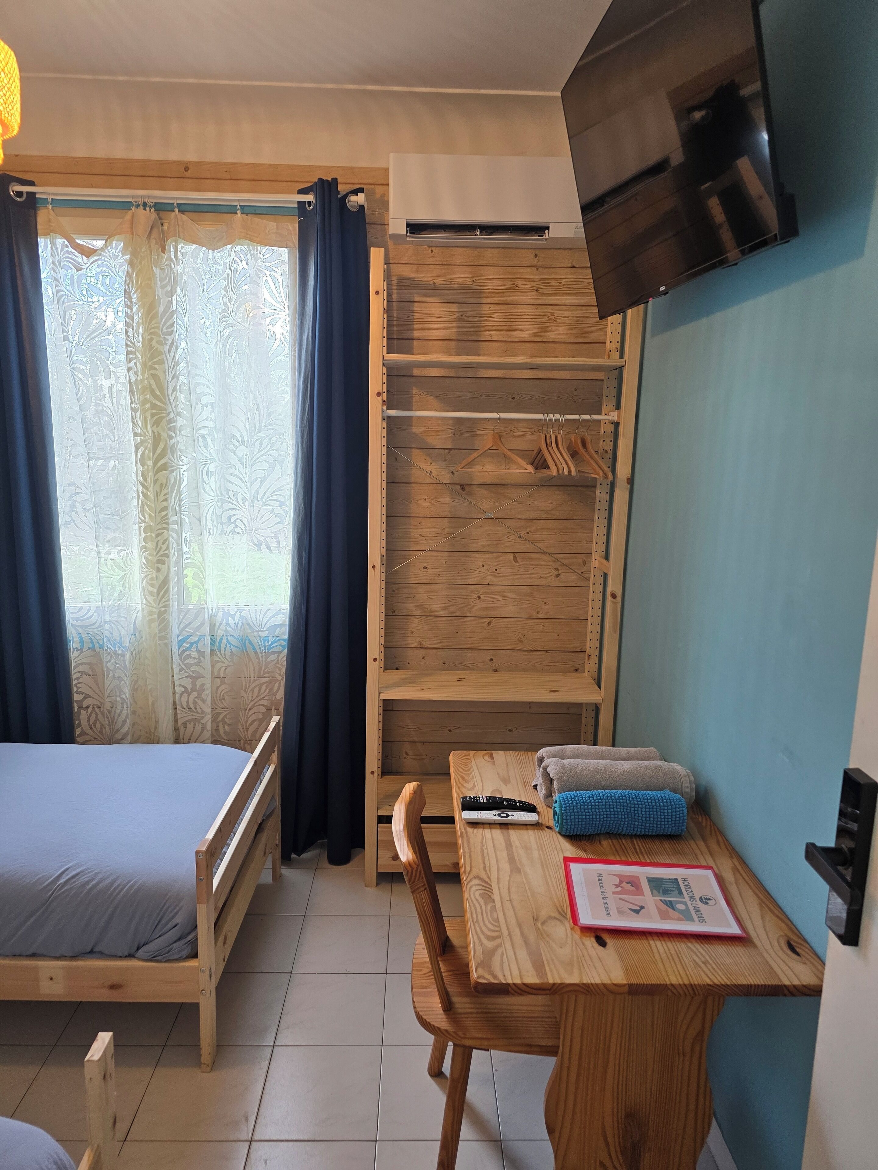 Una habitación con una cama, un escritorio de madera, una silla, un televisor montado en la pared y una ventana con cortinas.