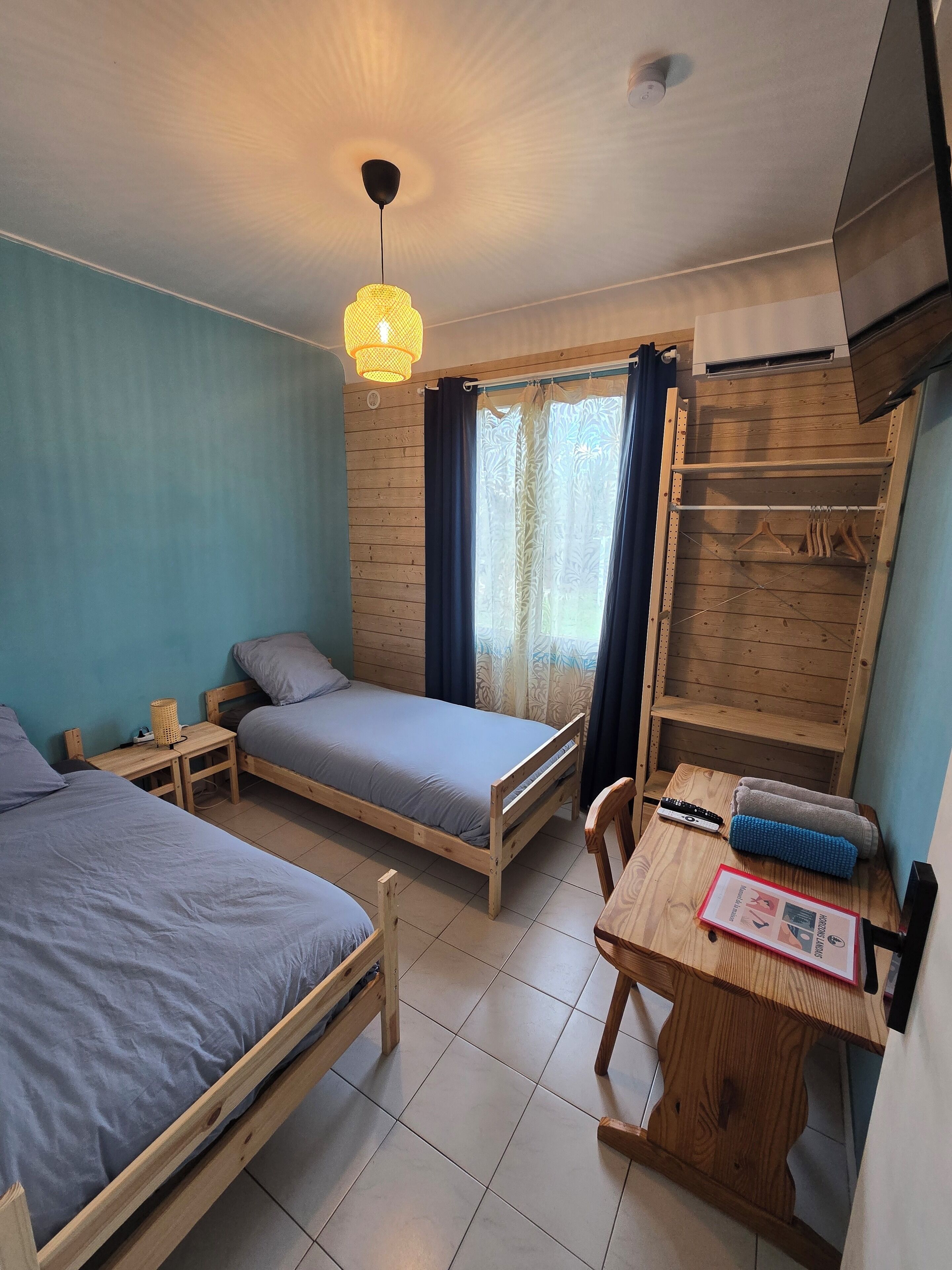 Une chambre à coucher avec deux lits en bois, un bureau avec une chaise, un luminaire suspendu et une armoire avec un miroir.