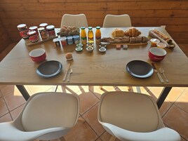 Una mesa de madera dispuesta para el desayuno, con pan, pastelería y bebidas, rodeada de sillas.