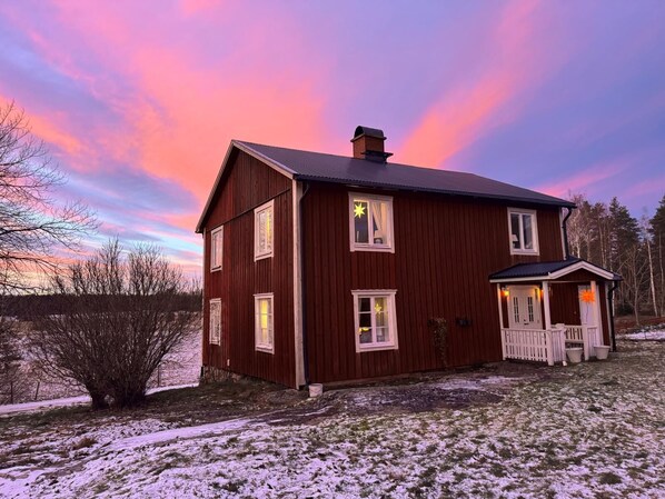 A two-story wooden house with a porch, surrounded by snow-covered ground and trees.