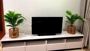 A flat-screen TV on a white entertainment unit with two potted plants, a painting on the wall, and a curtain. - Vortex Suites Klcc By Bukit Bintang (Kuala Lumpur)