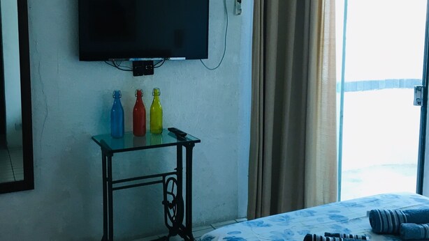Ein Hotelzimmer mit einem Bett, einem flachbildfernseher an der Wand, einem kleinen Tisch mit Wasserflaschen und einem Fenster mit Vorhängen.