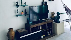 Ein Wohnzimmer mit einem Flachbildfernseher auf einem Holzboden, umgeben von Regalen mit Dekorationen, einer Hängematte und einer großen Vase.