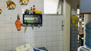 Eine Küche mit weiß gefliesten Wänden, einer Uhr und einem Fenster. Ein Tisch mit Tischdecke und zwei Hocker sind ebenfalls vorhanden.