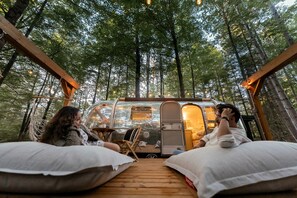 Un camping-car est garé dans une forêt, avec une terrasse en bois et deux personnes se détendent sur des coussins.