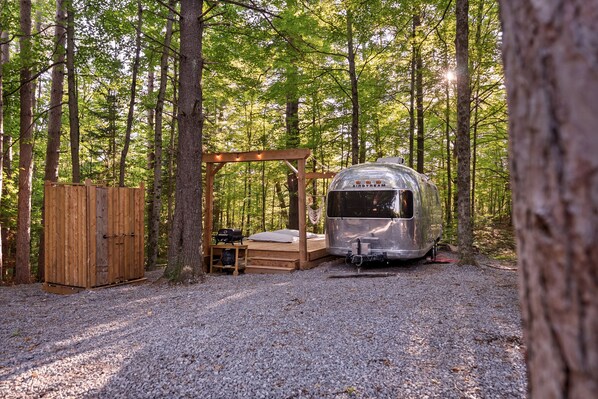 A forest campsite with a metal trailer, a wooden deck, and a small shed.
