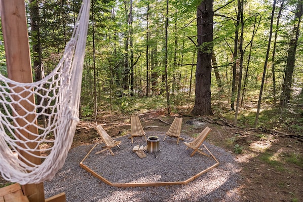 A forest clearing with a hammock, four wooden chairs, a fire pit, and a metal pot. - Airstream in Pines • Luxe Deck + Firepit + Shower (Burnstown)