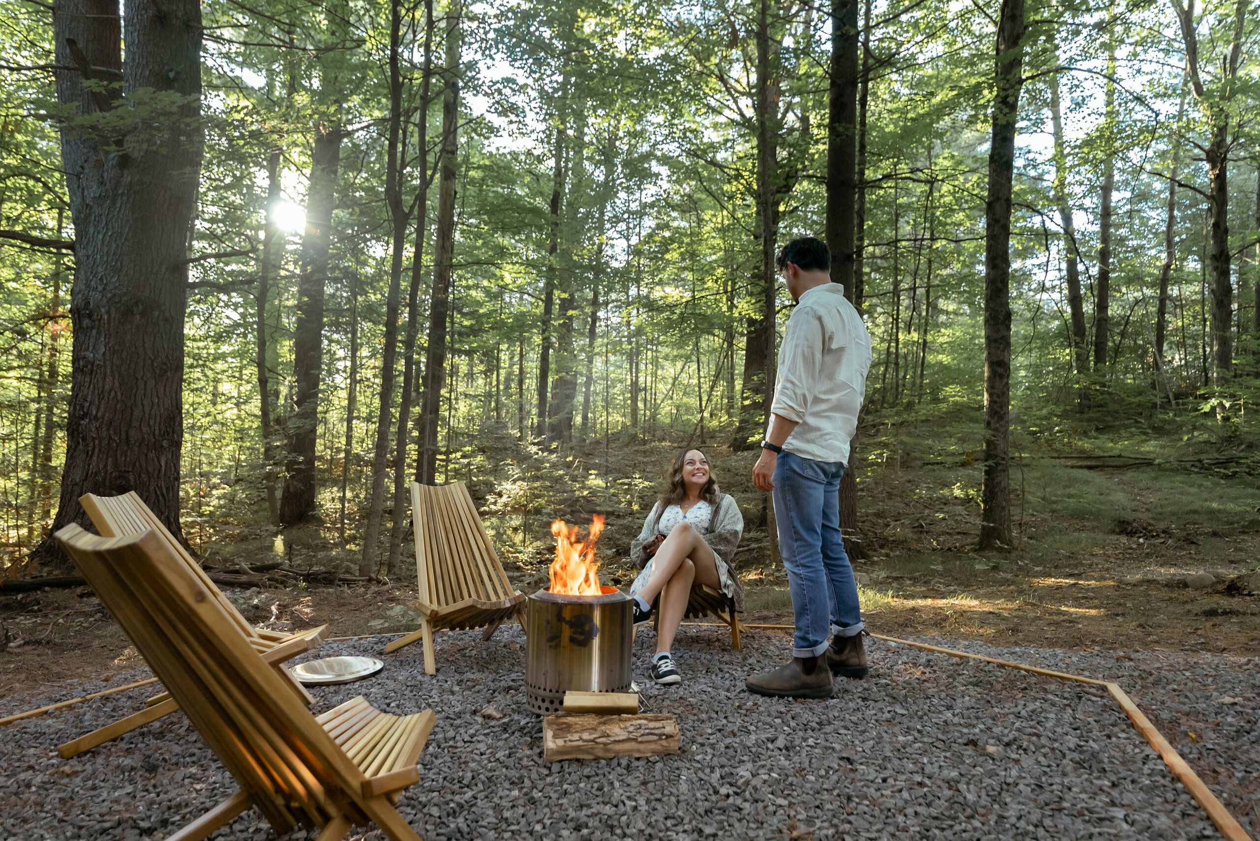 Campa de bosque con un hoyo para la fogata, dos sillas de madera y una persona de pie.