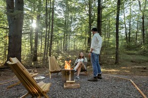 A forest campsite with a fire pit, two wooden chairs, and a person standing.
