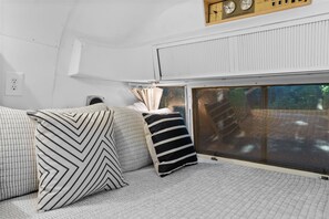 A cozy interior with a bed, pillows, a window, and a control panel. - Airstream in Pines • Luxe Deck + Firepit + Shower (Burnstown)