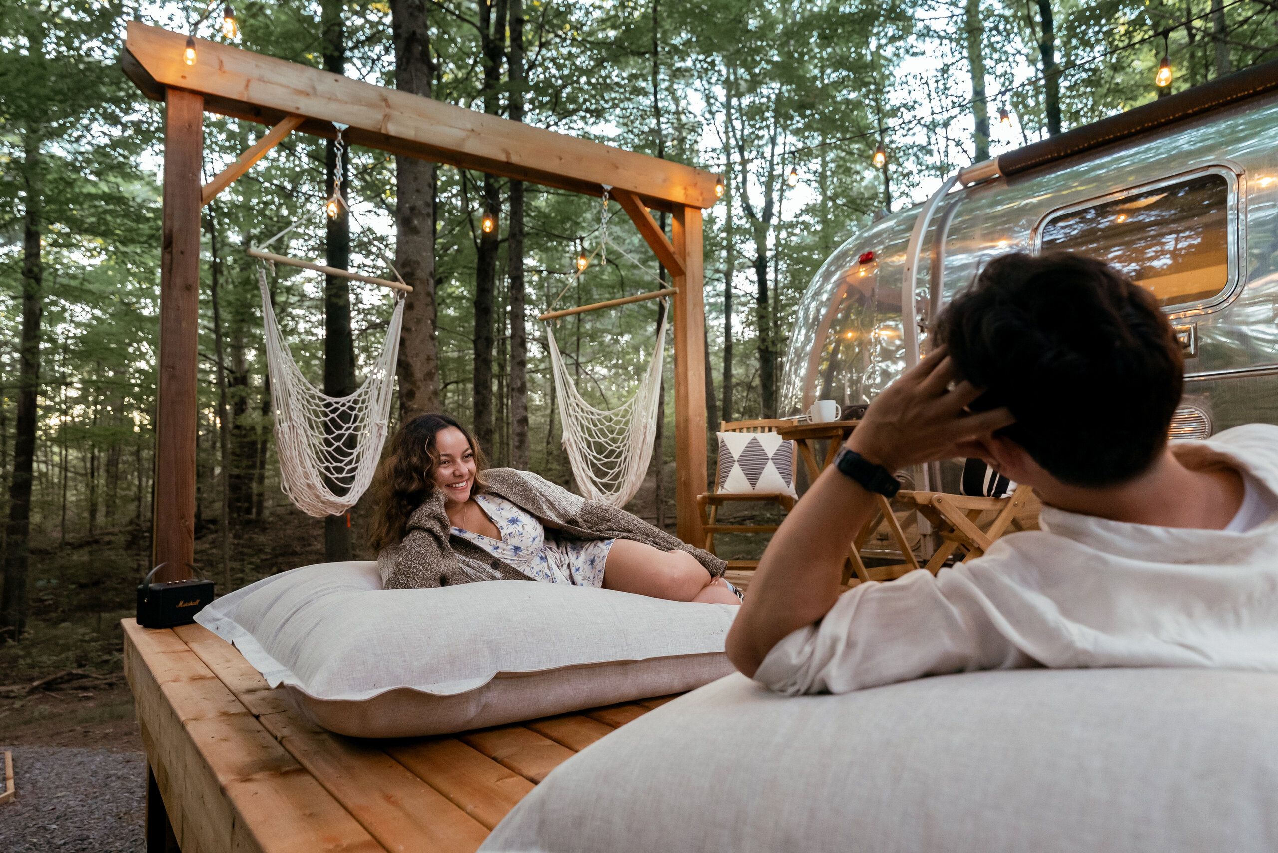 Una hamaca colgada de una estructura de madera en un bosque, una caravana estacionada cerca y una persona sentada en un banco.